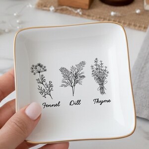 May include: A white square trinket dish with gold trim, featuring black line drawings of fennel, dill, and thyme, with their names below. The dish is held in a hand, with a marble surface and flowers in the background.