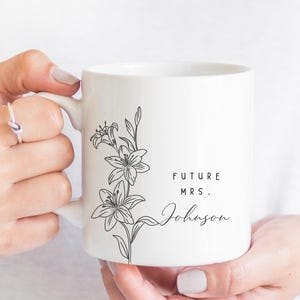 May include: White ceramic mug featuring a black floral design and the text "FUTURE MRS. Johnson". The mug has a handle and is held by a person. The design includes a detailed illustration of lilies.