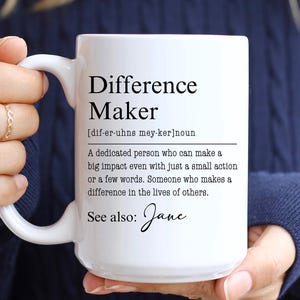 May include: White ceramic mug with the text "Difference Maker" and its definition. The mug also includes the phrase "See also: Jane". The mug is held by a person wearing a blue jumper.