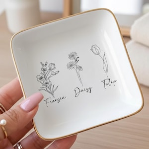 May include: A white square trinket dish with a gold rim, featuring delicate black line drawings of flowers: Freesia, Daisy, and Tulip. The dish is held in a hand, showcasing its elegant design. A perfect gift for a loved one.