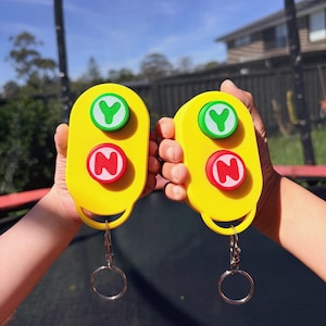May include: Two yellow, oval-shaped plastic keychains, each featuring two circular buttons. The top button is green with a white letter Y, and the bottom button is red with a white letter N. Each keychain includes a silver metal split ring attachment.
