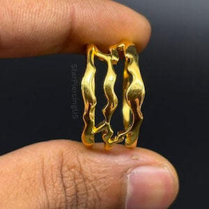 May include: A gold-colored ring featuring a triple-band design with wavy, organic edges. The surface of the metal is smooth and reflective. The text StarPiercingUS is printed vertically along the side of the ring structure.