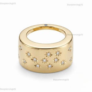 May include: A gold-colored ring with a wide band. The ring is adorned with multiple star-shaped designs, each containing a small, sparkling stone. A small blue stone is set on the top of the ring.