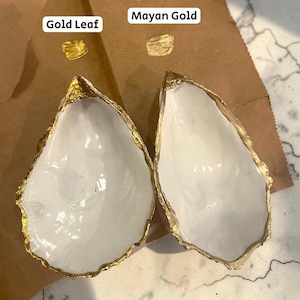 Puede incluir: Dos conchas de ostra blancas con borde de pan de oro. Los textos "Gold Leaf" y "Mayan Gold" est&aacute;n encima de peque&ntilde;as muestras de oro. Las conchas est&aacute;n sobre una superficie de papel marr&oacute;n.