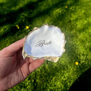 May include: A white oyster shell ring dish with gold-painted edges. The word "Bride" is written in elegant script across the center. The dish is held against a backdrop of green grass.