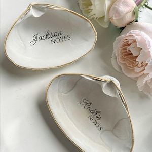 May include: Two white seashell-shaped trinket dishes with gold trim. One dish has the name "Jackson NOYES" in black script, and the other has "Ruthie NOYES". Soft pink and white roses are in the background.