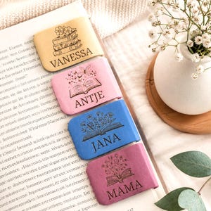 Könnte beinhalten: Vier rechteckige magnetische Lesezeichen in Gelb, Rosa, Blau und Lila. Jedes zeigt eine schwarze Gravur eines offenen Buches mit Blumenmotiven und einen Namen: Vanessa, Antje, Jana und Mama. Die Lesezeichen liegen auf einem aufgeschlagenen Buch.