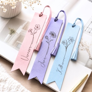 Könnte beinhalten: Drei rechteckige Lesezeichen in Pastellrosa, Lavendel und Hellblau, jeweils mit einer schwarzen floralen Strichzeichnung und einem personalisierten Namen in Schreibschrift. Jedes Lesezeichen hat eine passende Lederquaste am oberen Ende.