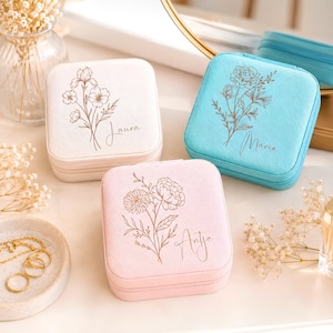 Könnte beinhalten: Drei quadratische Schmuckkästchen in Creme, Hellblau und Zartrosa, jeweils mit einer individuellen Blumen-Strichzeichnung und einem in Schreibschrift gedruckten Namen: Laura, Maria und Antje. Die Kästchen haben eine strukturierte Stoffoberfläche.