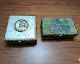 May include: Two antique decorative boxes. One box has a white, textured lid with a central, round, gold-framed design. The other box features a painted scene of a couple. Both boxes have a gold-colored metal frame.