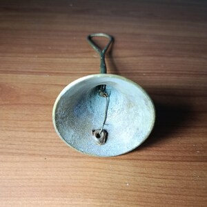 May include: A small, antique brass bell with a loop handle. The bell has a slightly tarnished appearance, with a visible clapper inside. The handle is a darker color than the bell itself. The bell is likely used for decorative purposes.
