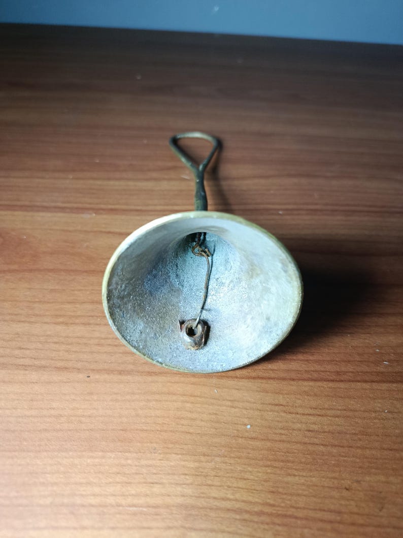 May include: A small, antique brass bell with a loop handle. The bell has a slightly tarnished appearance, with a visible clapper inside. The handle is a darker color than the bell itself. The bell is likely used for decorative purposes.