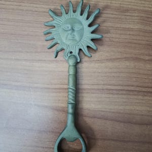 May include: A green metal bottle opener with a sun face design. The sun has a detailed face with rays extending outward. The handle is textured and leads to the bottle opener. The opener is on a wooden surface.