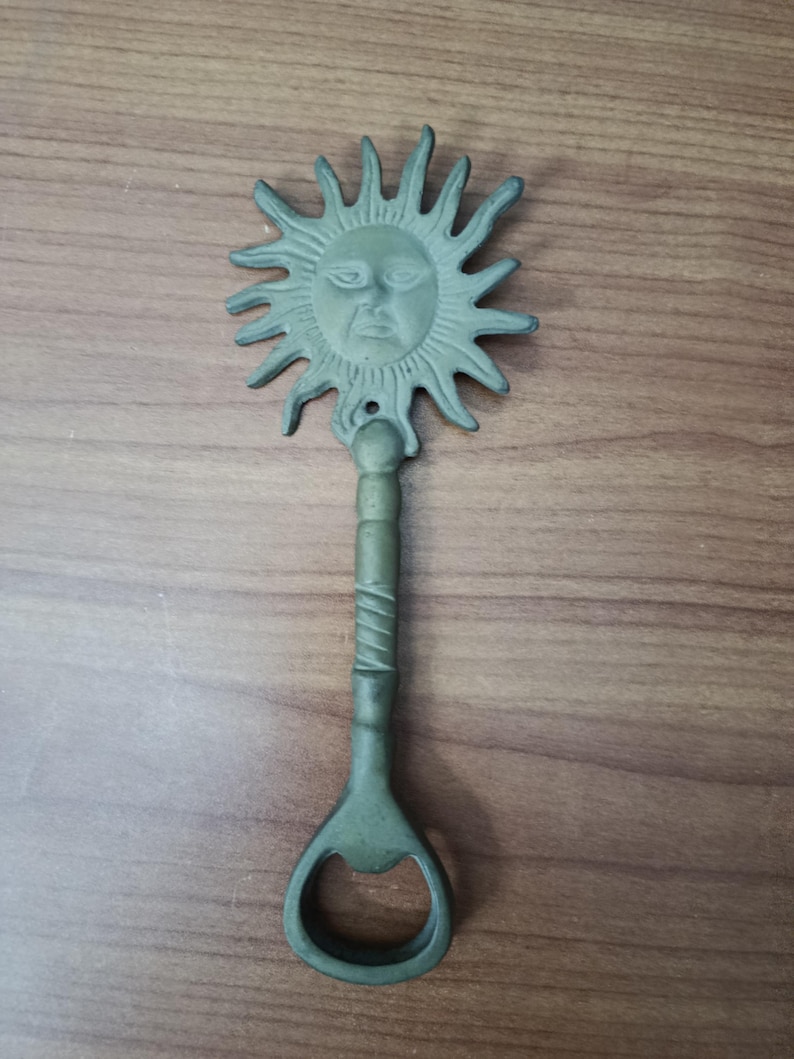 May include: A green metal bottle opener with a sun face design. The sun has a detailed face with rays extending outward. The handle is textured and leads to the bottle opener. The opener is on a wooden surface.