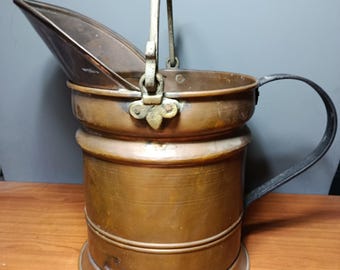 May include: A copper-toned metal coal scuttle with a hinged lid and a curved handle. The scuttle has a cylindrical body with a rounded base and a black metal handle. The lid is open, revealing the interior. The scuttle is likely an antique or vintage item.