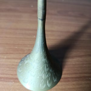 May include: A brass-colored, decorative candlestick holder with a flared base and a slender stem. The base features an etched feather design. The candlestick holder is displayed on a wooden surface.