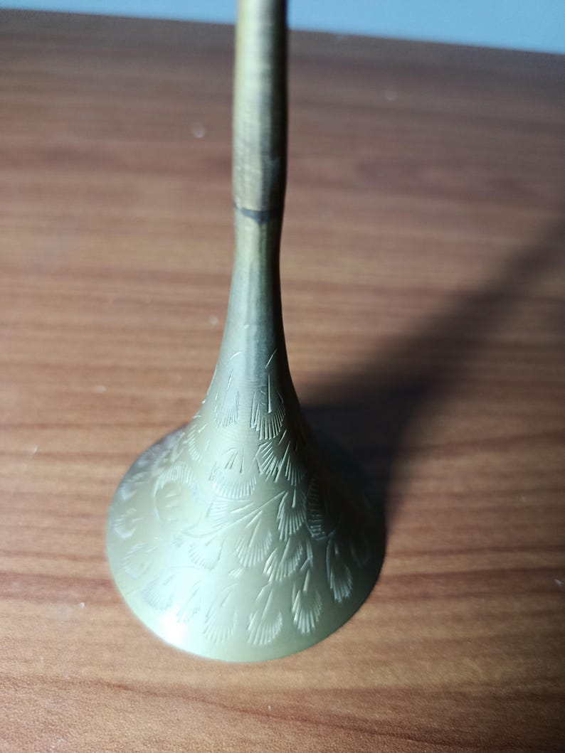 May include: A brass-colored, decorative candlestick holder with a flared base and a slender stem. The base features an etched feather design. The candlestick holder is displayed on a wooden surface.