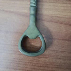 May include: A green bottle opener with a textured handle and a circular opening. The opener is made of metal and is designed for removing bottle caps. The handle has a series of ridges for grip.