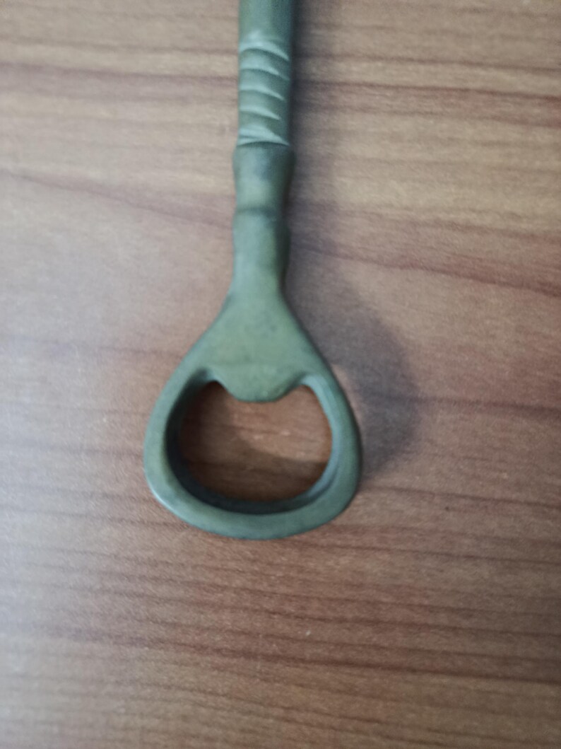 May include: A green bottle opener with a textured handle and a circular opening. The opener is made of metal and is designed for removing bottle caps. The handle has a series of ridges for grip.