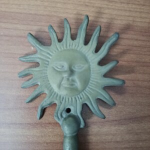 May include: A cast metal sun-shaped wall hook with a face in the center and radiating rays. The hook is painted in a muted green color and has a small hole for hanging.