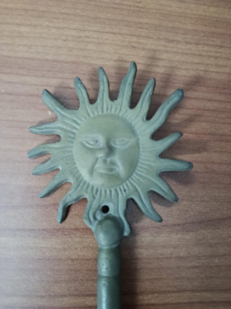 May include: A cast metal sun-shaped wall hook with a face in the center and radiating rays. The hook is painted in a muted green color and has a small hole for hanging.
