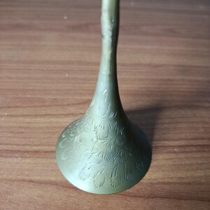 May include: A brass-colored, decorative candlestick holder with a flared base and a slender stem. The base features an etched floral pattern. The candlestick holder is displayed on a wooden surface.