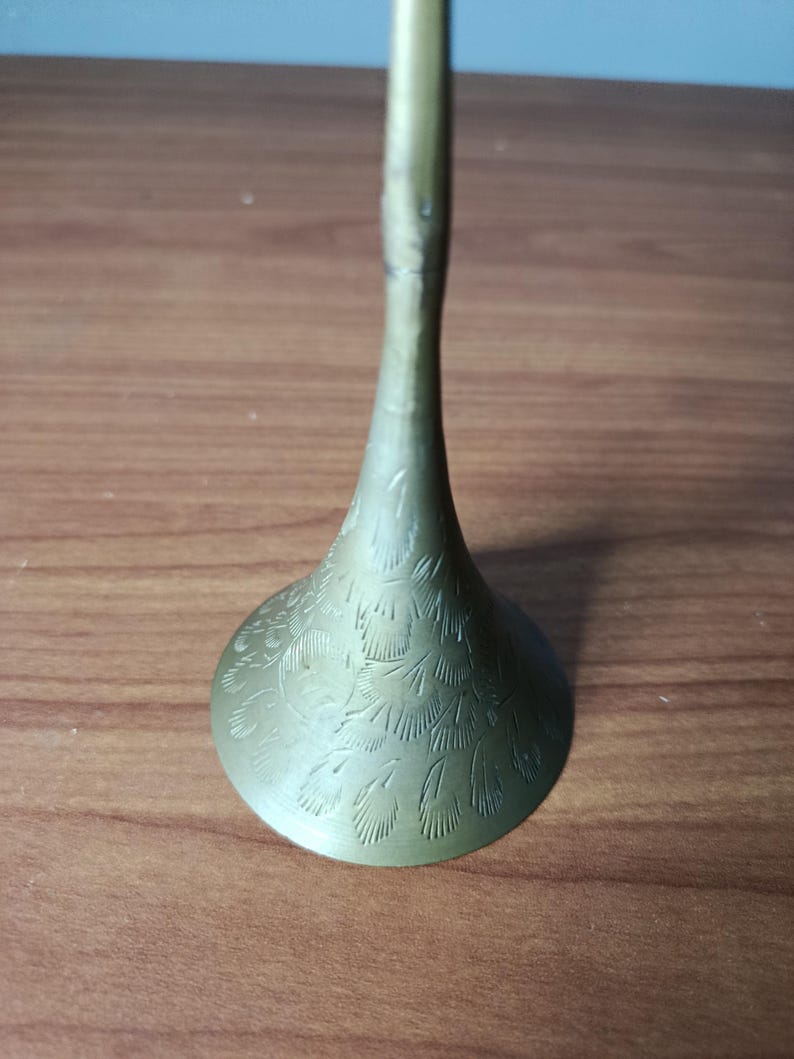 May include: A brass-colored, decorative candlestick holder with a flared base and a slender stem. The base features an etched floral pattern. The candlestick holder is displayed on a wooden surface.