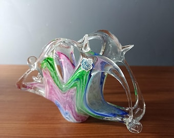 May include: A clear glass sculpture with a swirling design of pink, green, and blue. The art glass piece has a unique shape with curved lines and a small, attached label. The sculpture is displayed on a wooden surface.