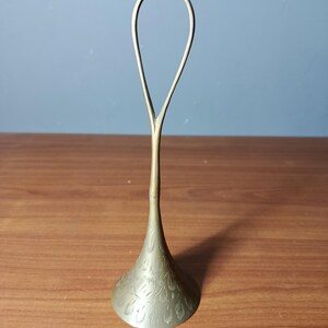 May include: A brass-colored, decorative object with a flared base and a looped handle. The base features an etched floral pattern. The object stands on a wooden surface against a blue wall.
