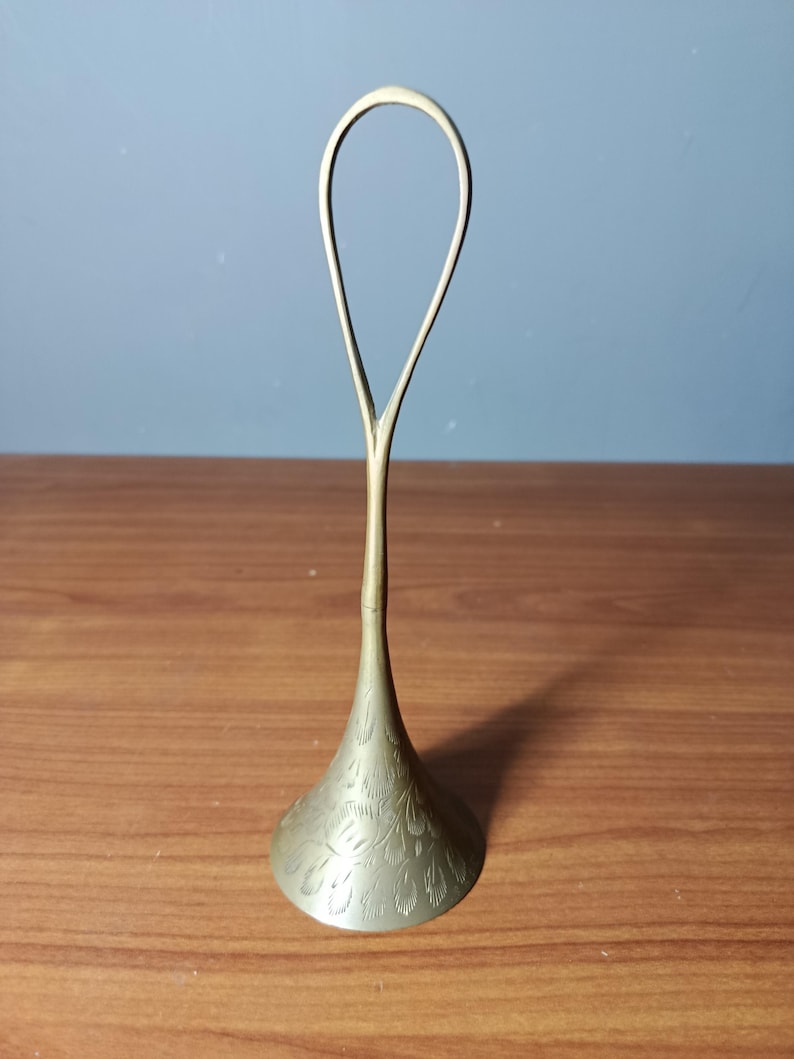 May include: A brass-colored, decorative object with a flared base and a looped handle. The base features an etched floral pattern. The object stands on a wooden surface against a blue wall.