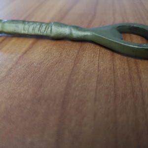 May include: An olive green metal bottle opener with a loop at one end and a textured handle. The opener is lying on a wooden surface, showcasing its design and color.