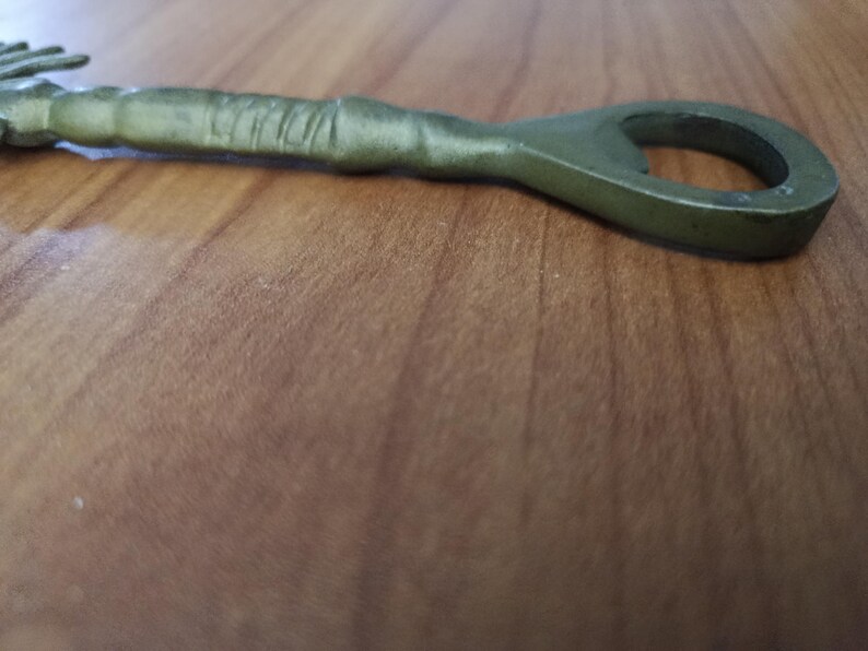 May include: An olive green metal bottle opener with a loop at one end and a textured handle. The opener is lying on a wooden surface, showcasing its design and color.