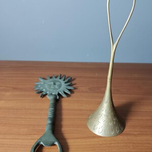 May include: A bronze bottle opener with a sun face design and a brass decorative stand with a floral pattern. The bottle opener is on the left, and the stand is on the right, both on a wooden surface.