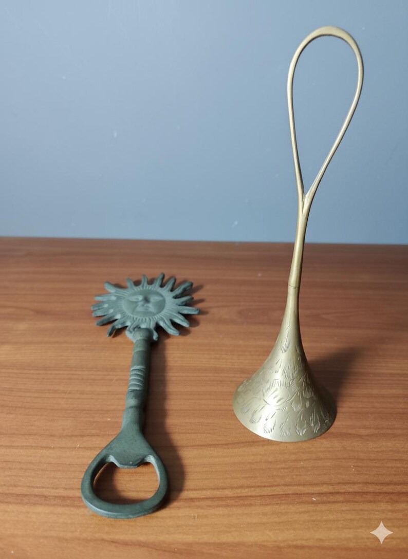 May include: A bronze bottle opener with a sun face design and a brass decorative stand with a floral pattern. The bottle opener is on the left, and the stand is on the right, both on a wooden surface.
