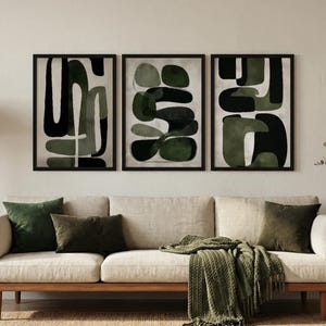 May include: Three framed abstract art prints with black frames, featuring olive green and black shapes. The prints hang above a beige sofa with green cushions and a green knitted throw. A wooden lamp and vase are also visible.
