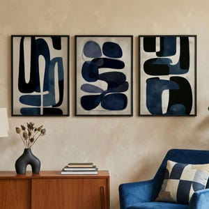 Puede incluir: Tres impresiones artísticas abstractas enmarcadas con formas azul marino y negras sobre un fondo claro. La obra de arte cuelga sobre un mueble de madera con una lámpara azul y un jarrón negro. A la derecha hay un sillón azul con un cojín estampado.