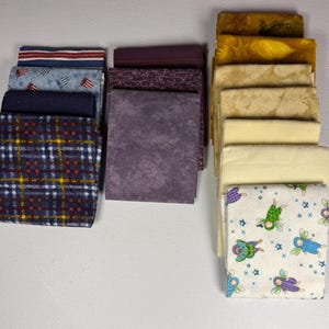 Puede incluir: Una colección de cuadrados de tela en varios colores y patrones. Las telas incluyen un estampado de cuadros escoceses, un estampado patriótico, un estampado floral y colores lisos en tonos de azul, morado y amarillo. Las telas están apiladas ordenadamente.