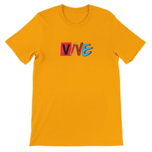May include: Mustard yellow t-shirt with the word "VIVE" in a colorful, retro design. The word "VIVE" is in a rectangular box with a pink background and black and white outlines. The letters "VE" are in blue.