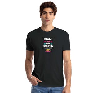 May include: A black short-sleeve t-shirt featuring a graphic print on the chest that reads MIAMI PLAYS THE WORLD 2026 in white and pink text, accompanied by icons of palm trees, a soccer ball, and a sunset over the horizon in vibrant colors.
