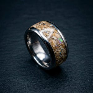 May include: A silver ring with a wide band. The centre of the ring is filled with a mosaic of tan and brown materials, with hints of iridescent colour. The ring is set against a dark background.