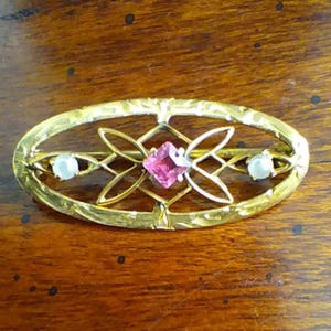 May include: Gold-toned oval brooch with an openwork design. The brooch features a central pink square gemstone, two small white pearls, and an intricate floral pattern. The brooch is set against a dark wood surface.