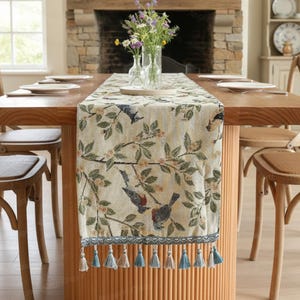 Könnte beinhalten: Beiger Tischläufer mit Vogel- und Blumenmuster, mit grünen Blättern und blauen Vögeln. Der Läufer hat einen blauen Rand mit Quasten. Der Läufer liegt auf einem Holztisch mit Stühlen.