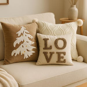 May include: A collection of decorative throw pillows in various designs. One pillow is tan with a white embroidered Christmas tree. Another is cream-colored with the word "LOVE" in brown textured letters. A third pillow has a cable knit design.