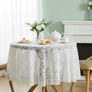 Puede incluir: Un mantel redondo de encaje blanco cubre una peque&ntilde;a mesa de madera, preparada para el t&eacute;. Una tetera blanca, una taza de t&eacute; y platos con pasteles est&aacute;n sobre la mesa. Un jarr&oacute;n de rosas blancas y vegetaci&oacute;n a&ntilde;ade un toque decorativo.