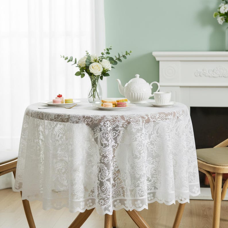 Puede incluir: Un mantel redondo de encaje blanco cubre una peque&ntilde;a mesa de madera, preparada para el t&eacute;. Una tetera blanca, una taza de t&eacute; y platos con pasteles est&aacute;n sobre la mesa. Un jarr&oacute;n de rosas blancas y vegetaci&oacute;n a&ntilde;ade un toque decorativo.