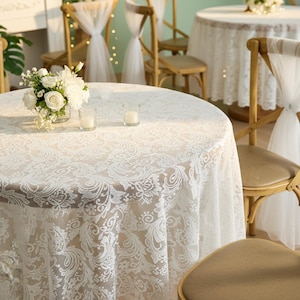 Puede incluir: Una mesa redonda cubierta con un mantel de encaje blanco con un dise&ntilde;o floral. Dos velas peque&ntilde;as y un jarr&oacute;n con flores blancas est&aacute;n sobre la mesa. Sillas de madera con adornos de tul blanco se ven en el fondo.