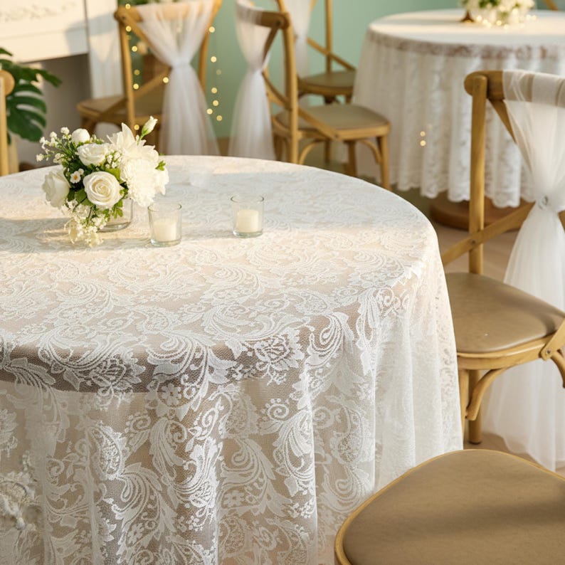 Puede incluir: Una mesa redonda cubierta con un mantel de encaje blanco con un dise&ntilde;o floral. Dos velas peque&ntilde;as y un jarr&oacute;n con flores blancas est&aacute;n sobre la mesa. Sillas de madera con adornos de tul blanco se ven en el fondo.