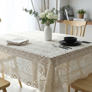 May include: A cream lace tablecloth, with a detailed openwork pattern and scalloped edge, covers a square table. A white vase with white flowers is in the centre. Black plates, a bowl, and cutlery are set on the table.