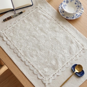 Op de afbeelding: Een rechthoekige witte kanten placemat met een bloemmotief en geschulpte randen. De placemat ligt op een beige linnen oppervlak. Een kop en schotel, een pen en een notitieboek zijn zichtbaar. Een gouden lepel rust op een blauw schaaltje.