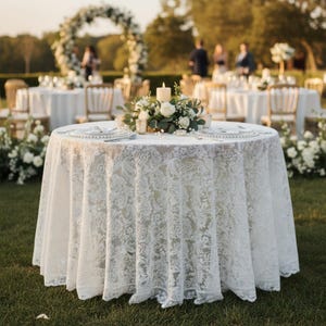 Puede incluir: Una mesa redonda preparada para un evento al aire libre, cubierta con un mantel de encaje blanco. La mesa está decorada con rosas blancas, vegetación, una vela encendida y cubiertos. Otras mesas y un arco floral son visibles en el fondo.